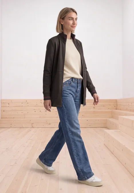 Jeune femme en veste noire, pull beige, jean bleu et baskets blanches marche dans un espace lumineux.