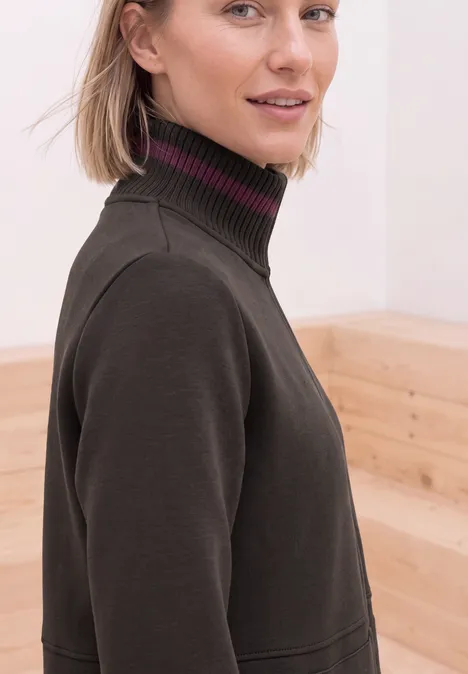 Femme blonde en pull col roulé foncé devant un fond en bois clair.