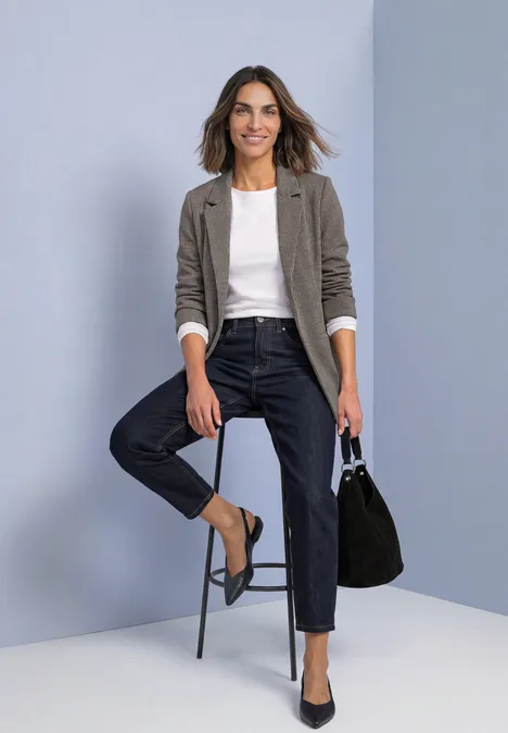 Vrouw in grijs colbert, wit T-shirt en donkerblauwe jeans, zittend op een kruk en met een zwarte handtas