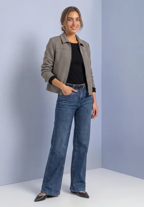 Vrouw in grijs kort jasje, zwart topje en wijde blauwe jeans tegen een lichte achtergrond