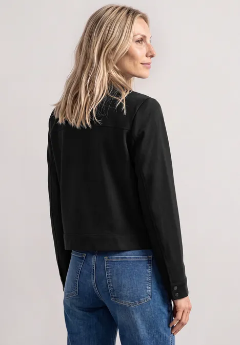 Jas met overhemdkraag en borstzakken Black Een vrouw met de rug naar de camera kijkt glimlachend naar rechts, draagt een zwart shirt en blauwe jeans.