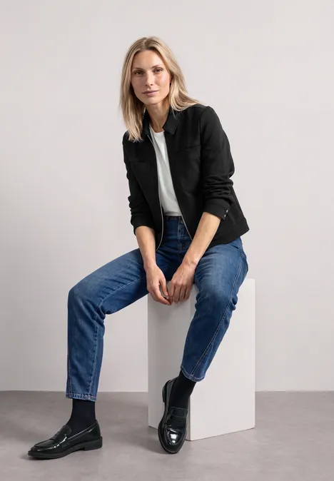 Jas met overhemdkraag en borstzakken Black Een blonde vrouw zit op een witte kubus, draagt een zwart colbert, wit t-shirt, blauwe jeans en zwarte laarzen.