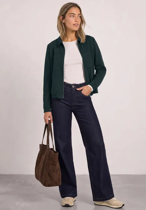 Jas met overhemdkraag en borstzakken stormy green Vrouw met wit topje, donker vest, wijde jeans, witte sneakers en bruine tas tegen neutrale achtergrond.