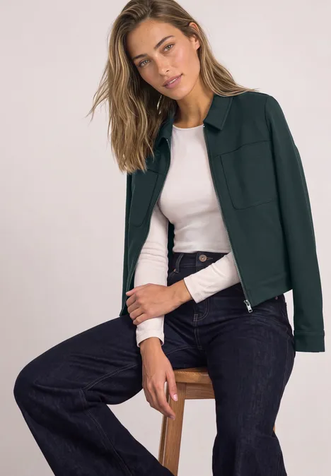 Jas met overhemdkraag en borstzakken stormy green Vrouw zit ontspannen op krukje, draagt donkergroene jas, licht shirt en zwarte jeans.
