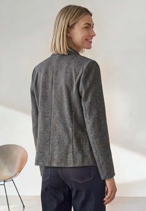 Frau mit blondem Bobhaarschnitt trägt grauen Blazer, steht im Profil, lächelt zur Seite.