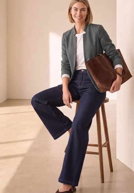 Eine Frau in weiten Jeans und Jacke sitzt auf einem Hocker, hält eine große braune Tasche.