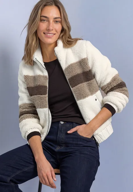 Teddy-Jacke im Colorblock-Look pure beige Frau sitzt lächelnd, trägt gestreifte, flauschige Fleecejacke in Creme- und Brauntönen, schwarzen Top und Jeans.