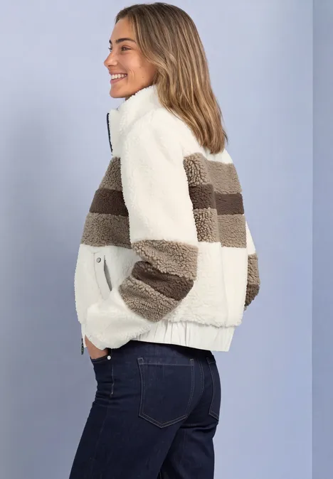 Teddy-Jacke im Colorblock-Look pure beige Junge Frau in cremeweißer Teddy-Jacke mit braunen Streifen und Jeans lächelt seitlich vor hellem Hintergrund.