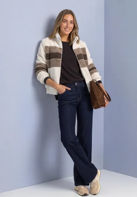 Teddy-Jacke im Colorblock-Look pure beige Frau mit gestreiftem Cardigan, dunkler Jeans, Sneakern und Tasche lehnt lächelnd vor lila Wand.