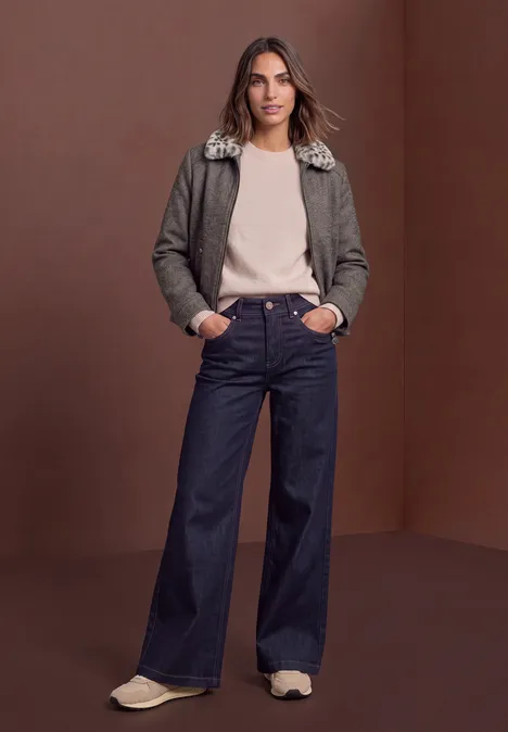 Vrouw in olijfgroen jasje met nepbontkraag, beige trui, wijde spijkerbroek en sneakers voor een bruine achtergrond.
