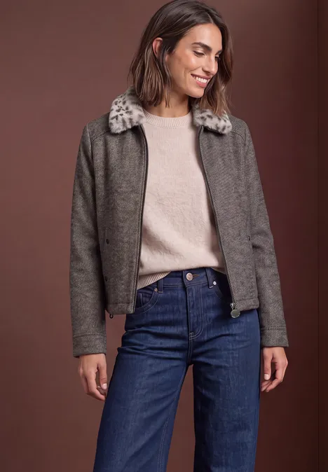 Vrouw met grijze jas met bontkraag, beige trui en spijkerbroek voor een bruine achtergrond.