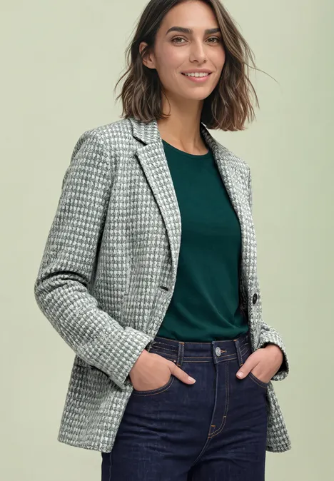 Vrouw met pied-de-poule blazer, groen shirt en spijkerbroek, handen in zakken, glimlachend.