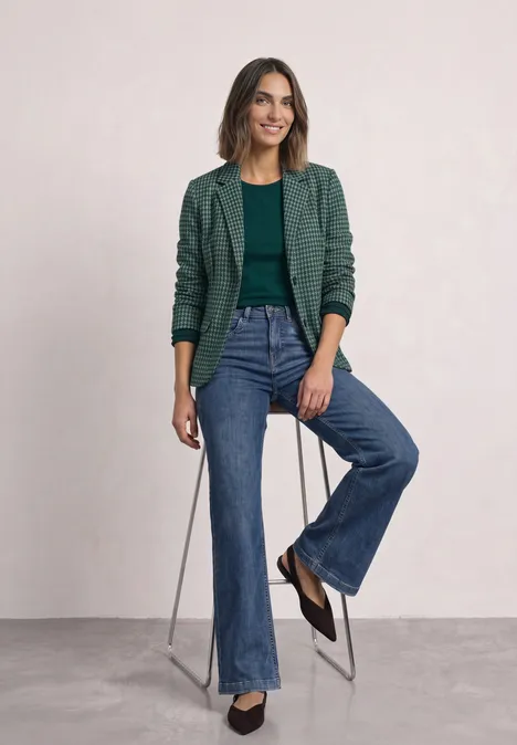 Basic Blazer mit Reverskragen und Muster stormy green Frau sitzt auf Hocker, trägt grünes Sakko, grünes Top, weite Jeans und schwarze Schuhe, lächelt.