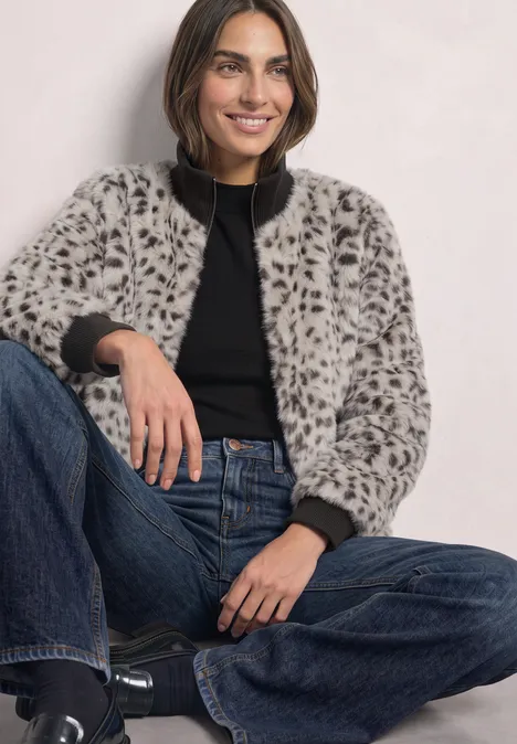 Fake Fur Jacke mit Animal Print black coffee Frau sitzt locker, trägt grauen Leo-Fleece, schwarzen Pullover und Jeans, lächelt entspannt.