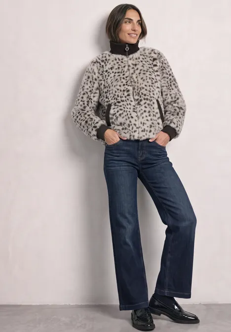 Fake Fur Jacke mit Animal Print black coffee Frau in Leo-Kunstpelzjacke, dunklen Jeans und schwarzen Schuhen lehnt an einer hellen Wand.
