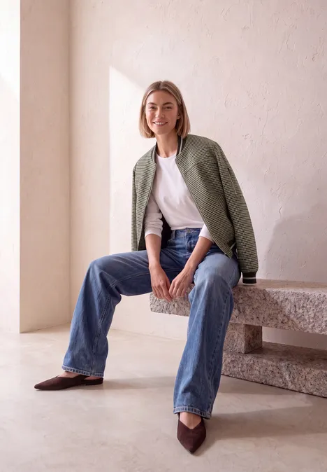 Blouson mit Zipper und Karomuster sanded amber Frau sitzt lässig auf einer Steinbank, trägt Jeans, weißes Shirt und grünes Sakko, lächelt in minimalistischem Raum.