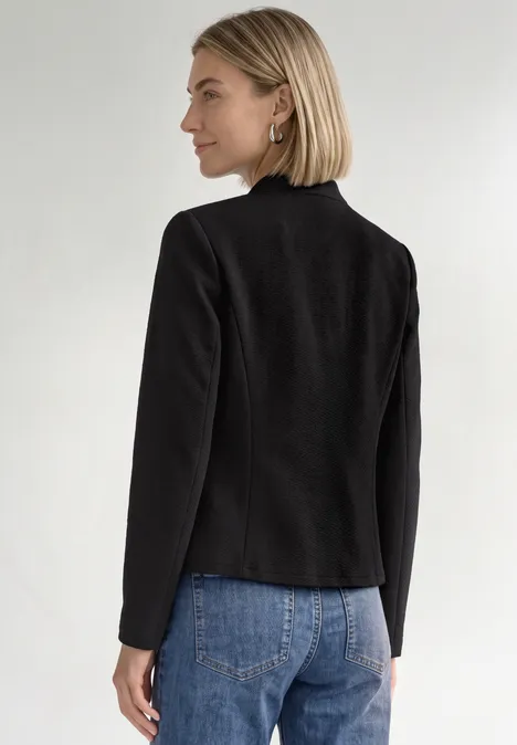 Frau mit kurzem blondem Haar in schwarzem Blazer und blauen Jeans, seitlich von hinten vor weißem Hintergrund.