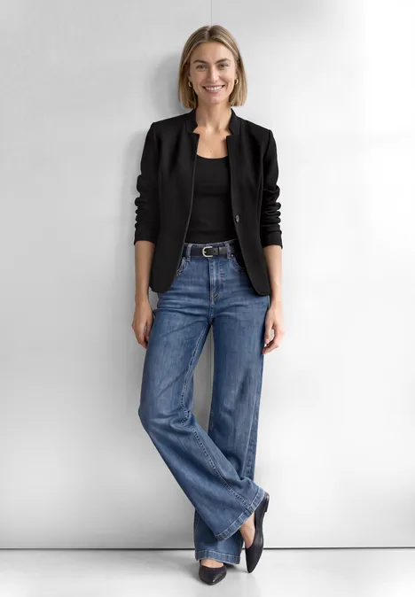 Eine Frau steht vor hellem Hintergrund in schwarzer Jacke, schwarzem Top, weitem Jeans und schwarzen Schuhen.