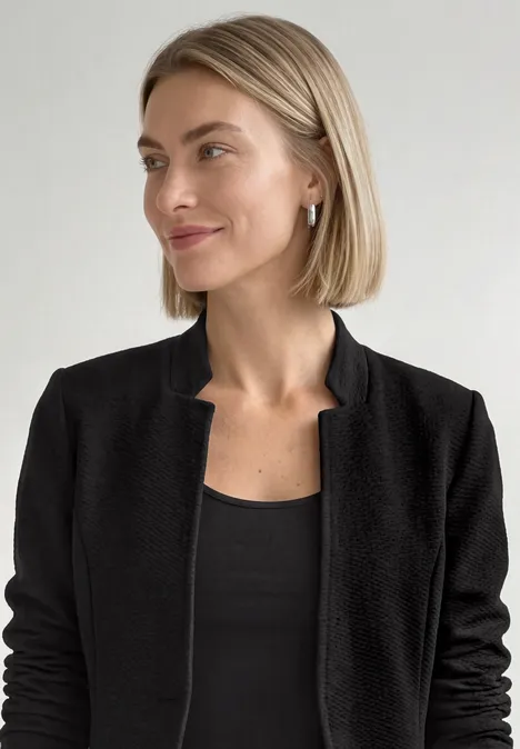 Eine Frau mit schulterlangen blonden Haaren trägt eine schwarze Jacke und lächelt leicht vor hellem Hintergrund.