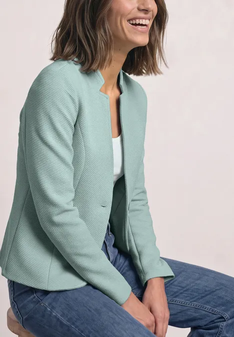 Een glimlachende vrouw zit met haar handen op haar schoot in een mintgroene blazer en jeans.