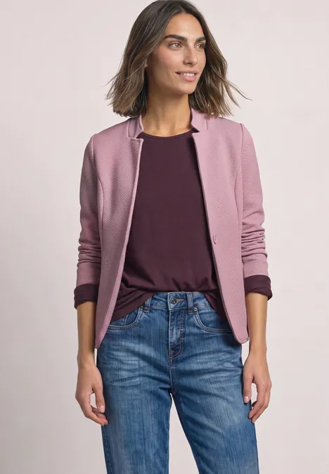 Frau in casual Outfit trägt rosa Blouson und Jeans vor hellem Hintergrund.