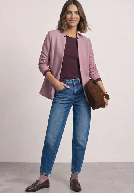 Eine Frau trägt ein rosa Sakko, burgunderfarbenes Shirt, blaue Jeans und Loafers und hält eine braune Clutch.