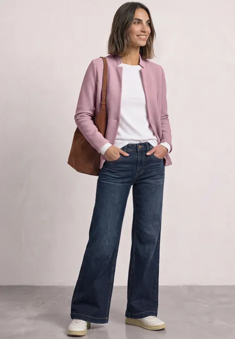 Frau in weitem Jeans, weißem Shirt, rosafarbenem Cardigan, brauner Tasche und Sneakern posiert lächelnd.