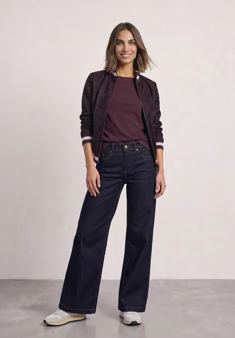 Frau in bordeauxroter Bluse, violetter Bomberjacke, weiten Jeans und weißen Sneakern.