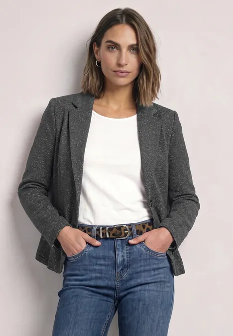 Vrouw in jeans, wit t-shirt en grijze blazer staat ontspannen met handen in de zakken.