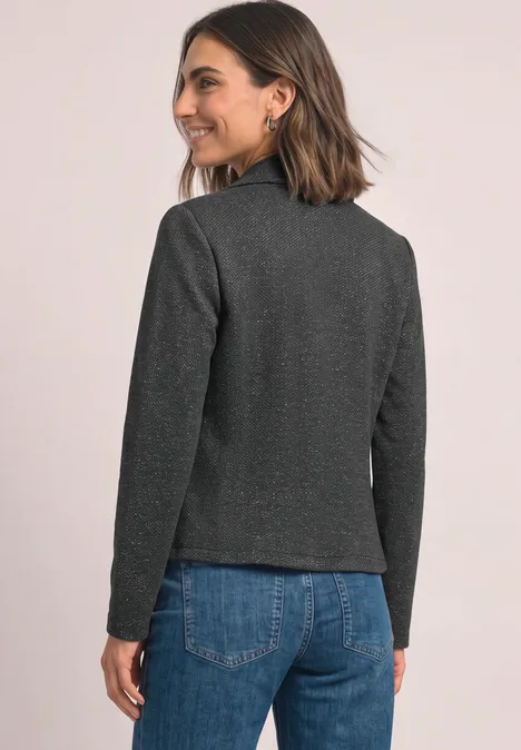 Vrouw in een grijs jasje en blauwe jeans, kijkt over haar schouder, glimlachend.