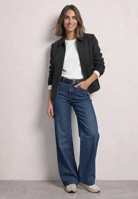 Femme en veste noire, tee-shirt blanc, jean large bleu et baskets blanches sur fond neutre.