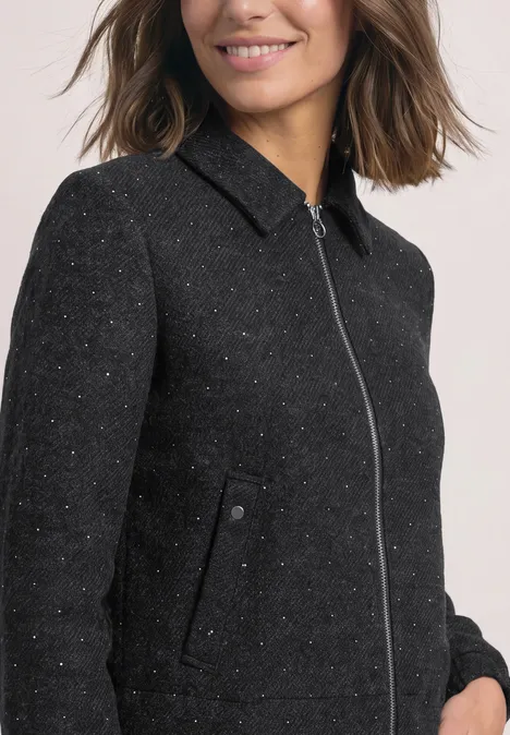 Une femme aux cheveux bruns porte une veste noire texturée à fermeture éclair et col montant.
