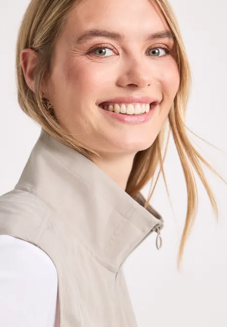 Een glimlachende vrouw met blond haar draagt een licht gilet voor een witte achtergrond.