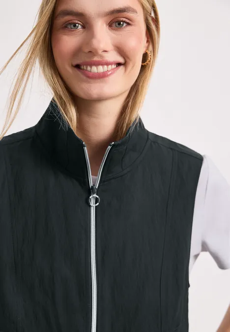Model met vervaagd gezicht draagt een zwarte mouwloze bodywarmer met rits en ringtrekker over een wit shirt.