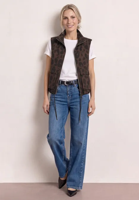 Femme en t-shirt blanc, gilet sans manches marron à motif, jean évasé bleu et chaussures noires.