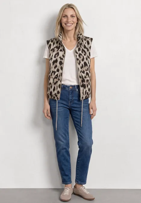 Frau trägt weißes T-Shirt, Leopardenweste, blaue Jeans und beige Sneaker vor hellem Hintergrund.