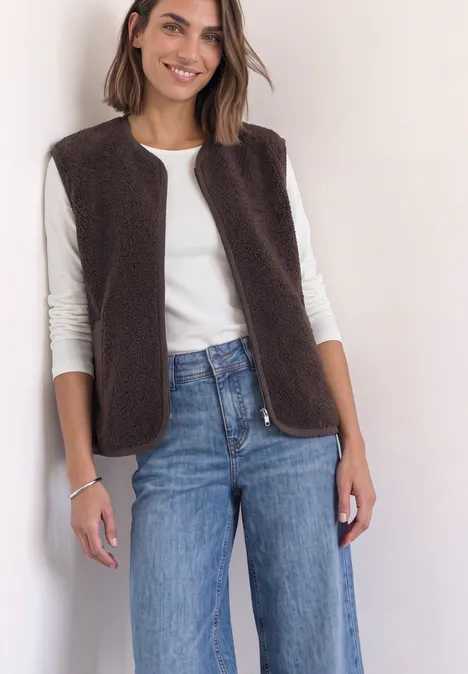 Femme souriante porte un haut blanc, une veste sans manches en polaire marron et un jean bleu.