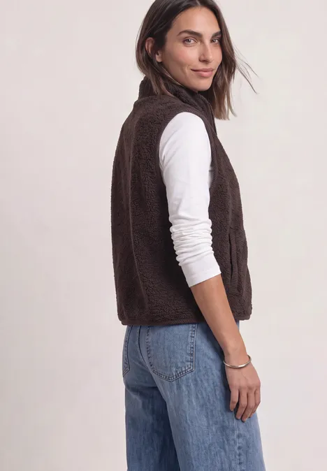 Femme en trois-quarts porte une veste en polaire marron, un t-shirt blanc à manches longues et un jean bleu sur fond clair.