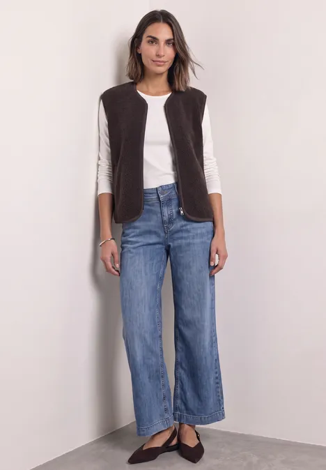 Une femme devant un mur clair porte un haut blanc, un gilet marron, un jean large et des ballerines noires.