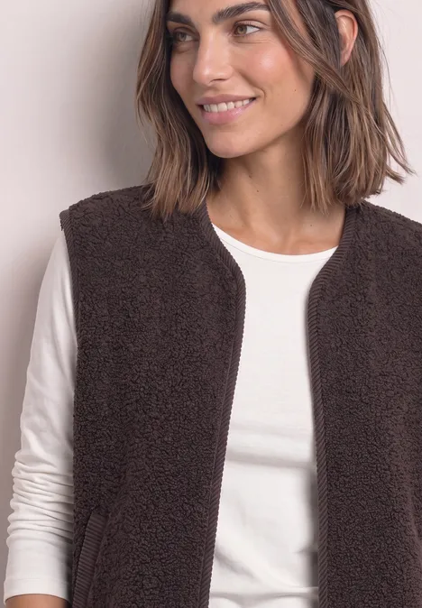 Une femme souriante porte une veste en polaire brune sur un t-shirt blanc à manches longues.