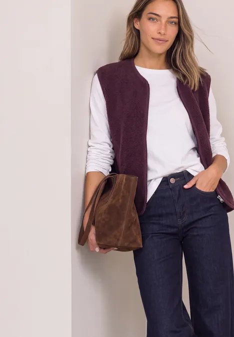 Frau in weißem Shirt, lila Weste und Jeans, trägt eine braune Wildledertasche vor hellem Hintergrund.