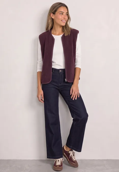 Frau in weißem Shirt, lila Weste, weiten Jeans und braunen Schuhen vor hellem Hintergrund, lächelnd.