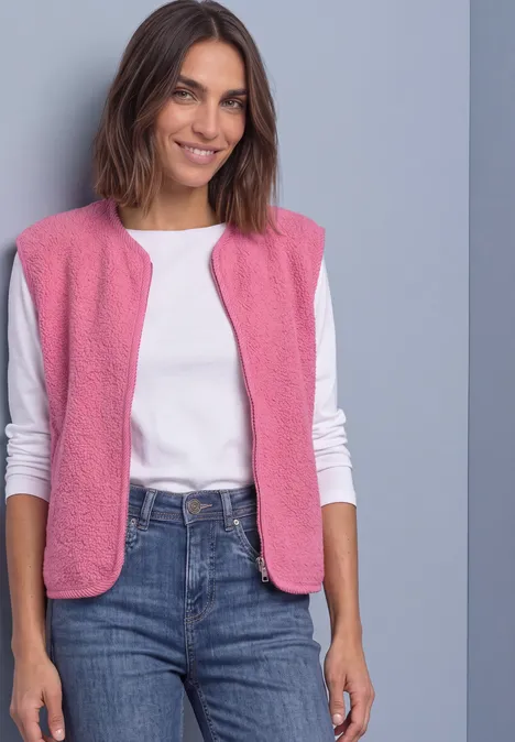 Eine lächelnde Frau in pink ärmelloser Weste, weißem Shirt und Jeans vor grauem Hintergrund.