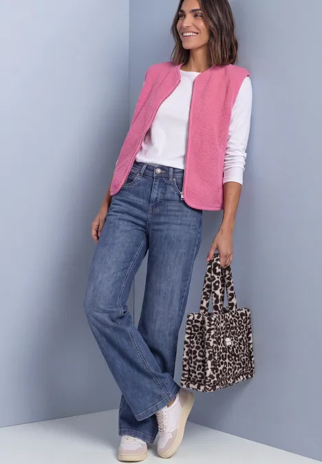 Frau in weißem Langarmshirt, pinker Weste und weiten Jeans, weiße Sneaker, hält gepunktete Tasche und lächelt.