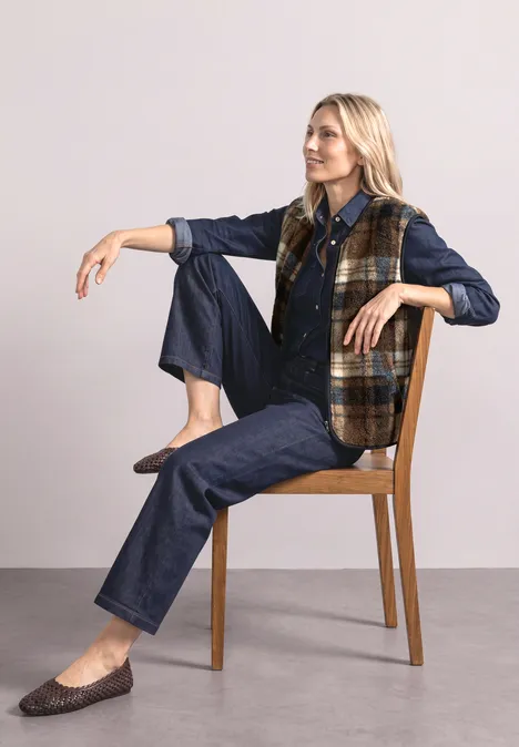 Blonde Frau in Jeans, kariertem Weste und Loafers sitzt entspannt auf einem Stuhl.