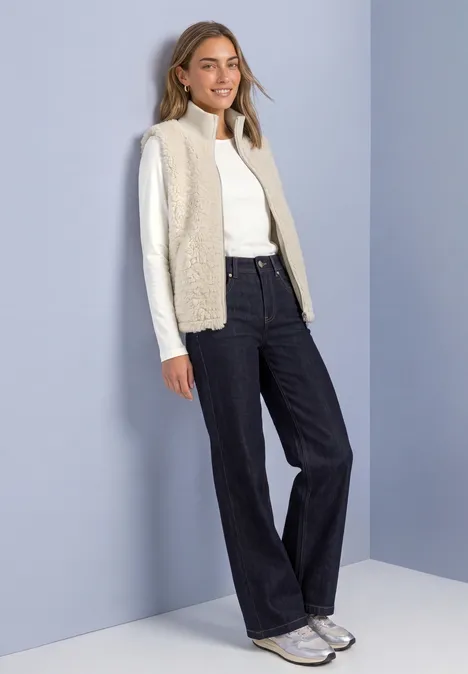 Frau mit hellem Rollkragenpullover, beige Weste, weiten Jeans und Sneakern vor pastellfarbenem Hintergrund.