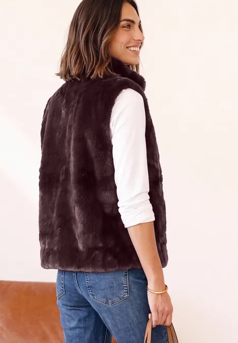Frau im Profil mit schwarzer Fellweste, weißem Langarmshirt und Jeans, lächelnd.