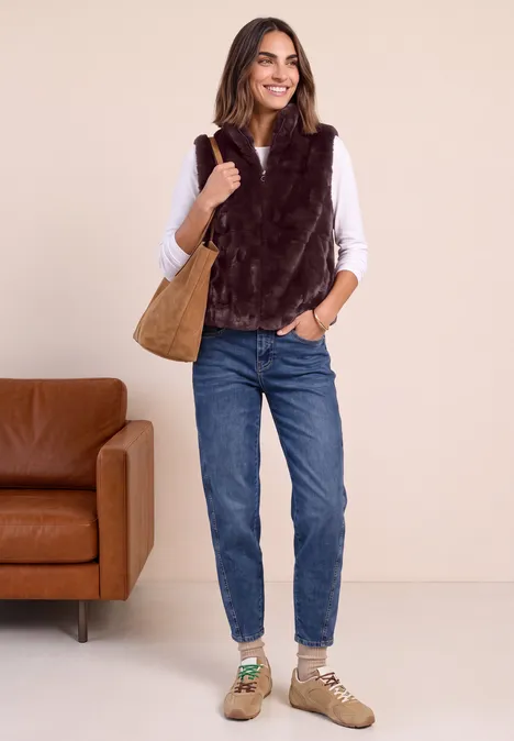 Junge Frau mit weißer Bluse, brauner Kunstfellweste, blauen Jeans, beigen Schuhen und brauner Handtasche.