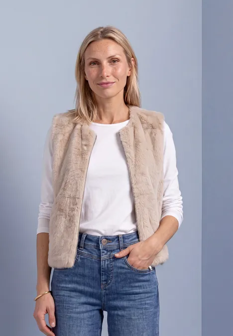 Eine Frau in weißem Shirt und beigem Fellweste, Jeans vor grauem Hintergrund.