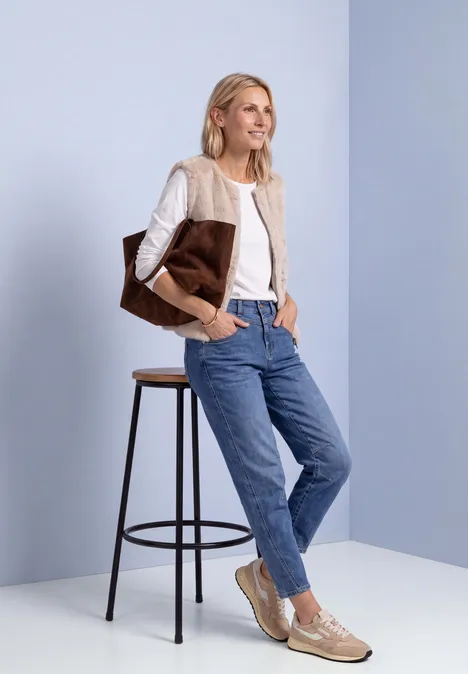 Eine lächelnde Frau in beigem Cardigan, weißem Shirt und Jeans lehnt an einem Hocker und trägt eine große Tasche.